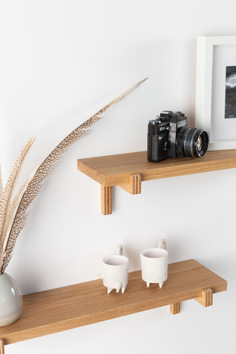 May include: Two wooden shelves with a vintage camera on the top shelf and two white ceramic mugs on the bottom shelf. The shelves are mounted on a white wall.