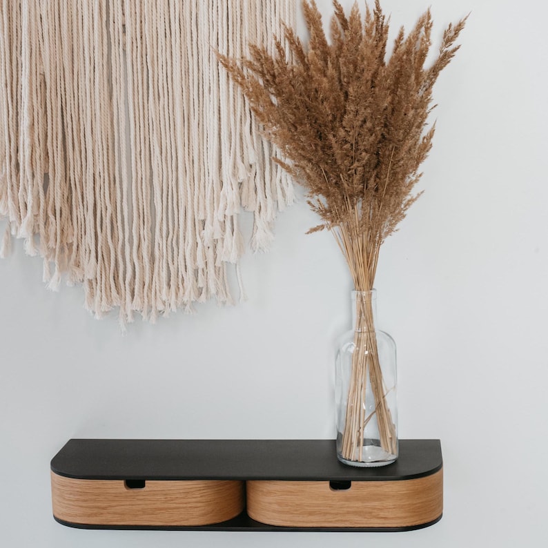 May include: A black floating shelf with two wooden drawers, mounted on a white wall. A clear glass vase with dried pampas grass sits on the shelf.