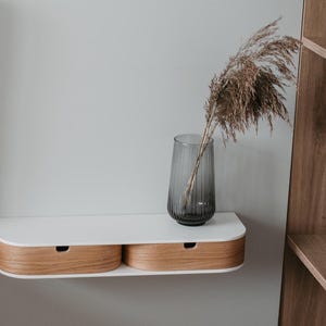 May include: A white floating shelf with two wooden drawers. The shelf is mounted on a white wall and has a gray glass vase with dried flowers on top.