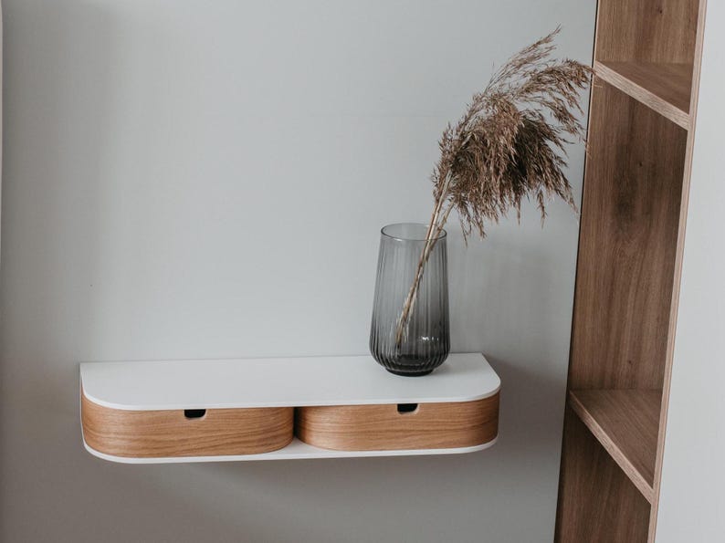 May include: A white floating shelf with two wooden drawers. The shelf is mounted on a white wall and has a gray glass vase with dried flowers on top.