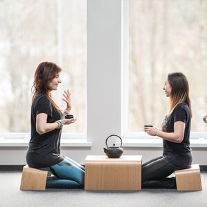 May include: Two people sit on wooden meditation cushions facing each other, holding small cups. A black teapot sits on a wooden table between them.