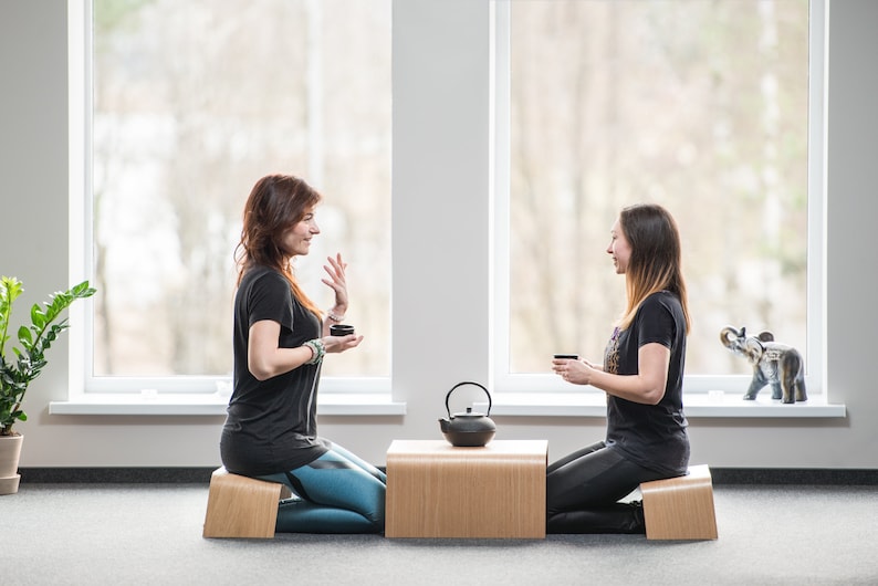 May include: Two people sit on wooden meditation cushions facing each other, holding small cups. A black teapot sits on a wooden table between them.