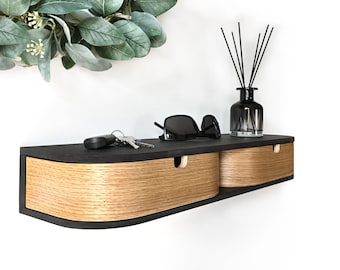 Può includere: Uno scaffale a muro nero con due cassetti. I cassetti sono in legno con una finitura marrone chiaro. Lo scaffale ha un bordo anteriore curvo ed è decorato con una bordura nera. Lo scaffale contiene un paio di occhiali da sole, un set di chiavi e una bottiglia di vetro con canne.