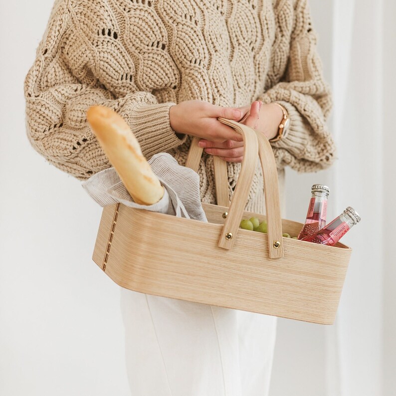 May include: A woman is holding a wooden picnic basket with a baguette, grapes, and bottles of pink beverage inside. The basket has two handles and is made of light brown wood.