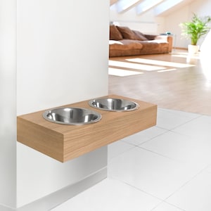 May include: A wooden pet food and water bowl stand mounted on a white wall. The stand has two stainless steel bowls.