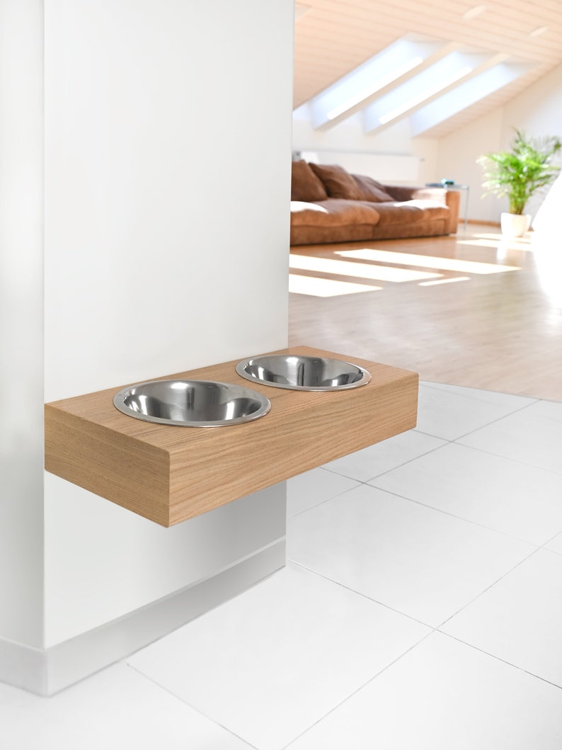 May include: A wooden pet food and water bowl stand mounted on a white wall. The stand has two stainless steel bowls.