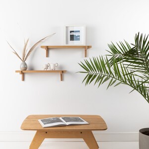 May include: Two wooden shelves with decorative brackets mounted on a white wall. The top shelf has a framed picture with a blue and white landscape. The bottom shelf has a vase with dried grasses and two white ceramic mugs. A wooden coffee table with a book on top sits in front of a potted palm plant.