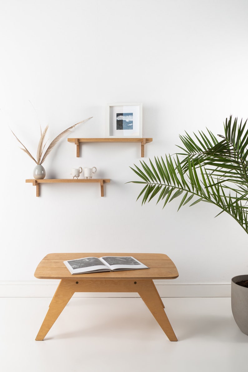 May include: Two wooden shelves with decorative brackets mounted on a white wall. The top shelf has a framed picture with a blue and white landscape. The bottom shelf has a vase with dried grasses and two white ceramic mugs. A wooden coffee table with a book on top sits in front of a potted palm plant.