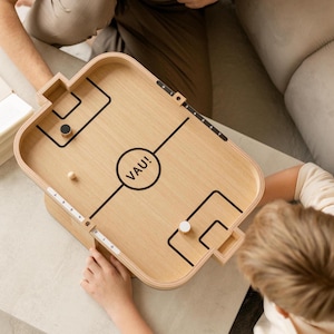 May include: A wooden tabletop game with a light wood finish. The game features black lines marking the play area, with the word "VAU!" in a circle. Includes a black and white puck and a small wooden ball. The game is rectangular with rounded corners.