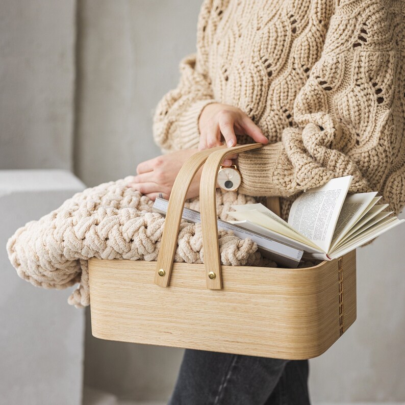 May include: A light brown knitted blanket is placed in a wooden basket with a natural finish. The basket has two handles and is filled with books.