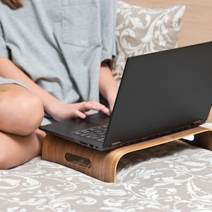 May include: A person is sitting on a bed with a laptop on a wooden stand. The laptop is open and the person is typing on the keyboard.