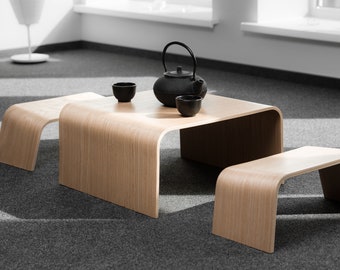 Puede incluir: Una mesa de café de madera con un borde curvo y dos taburetes de madera a juego. Una tetera negra y tres tazas de té negras están sobre la mesa.