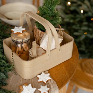 May include: A wooden basket with handles filled with Christmas cookies, a jar of cookies, and wrapped gifts. The basket is sitting on a wooden table with a white cloth and more cookies.