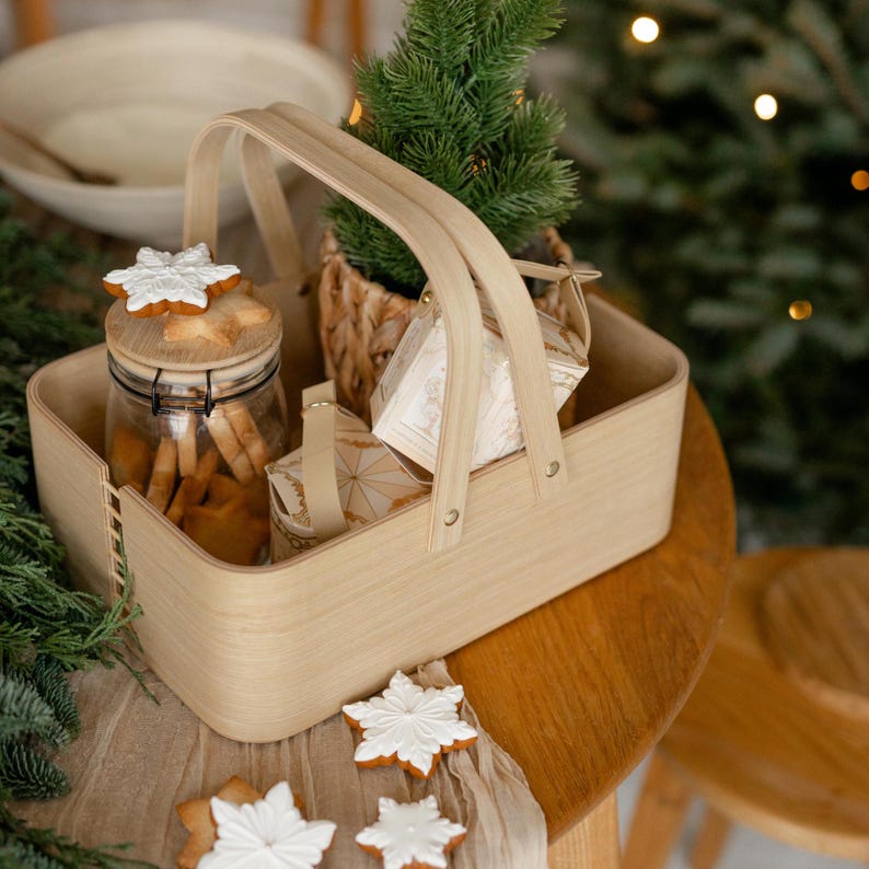 May include: A wooden basket with handles filled with Christmas cookies, a jar of cookies, and wrapped gifts. The basket is sitting on a wooden table with a white cloth and more cookies.