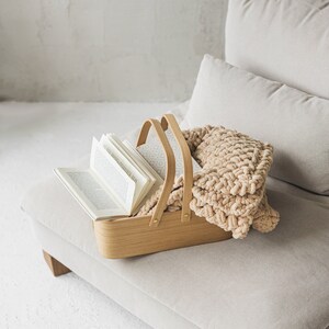 May include: A wooden basket with a book and a knitted throw blanket inside. The basket is sitting on a light beige couch.