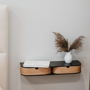 May include: A black floating shelf with two wooden drawers, mounted on a white wall. A white vase with dried brown grasses sits on the shelf.