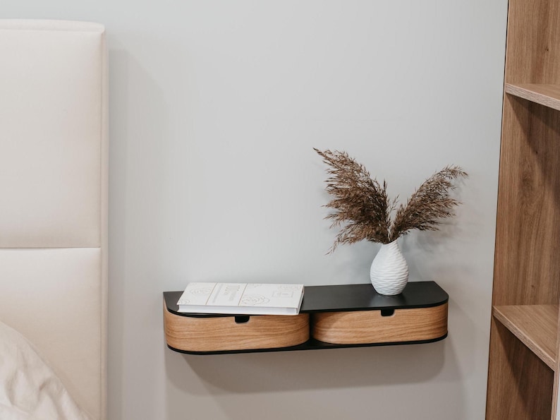 May include: A black floating shelf with two wooden drawers, mounted on a white wall. A white vase with dried brown grasses sits on the shelf.