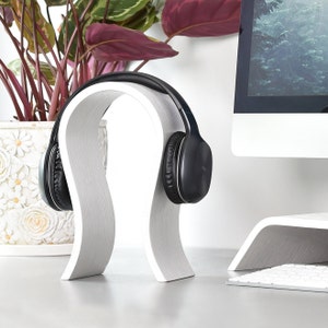 Könnte beinhalten: Weißer, gebogener Kopfhörerständer aus Holz. Der Ständer hält einen schwarzen Over-Ear-Kopfhörer.