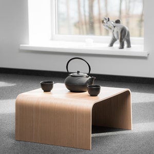 May include: A wooden table with a curved edge, featuring a black cast iron teapot and two small ceramic cups. The table is set on a gray carpet.