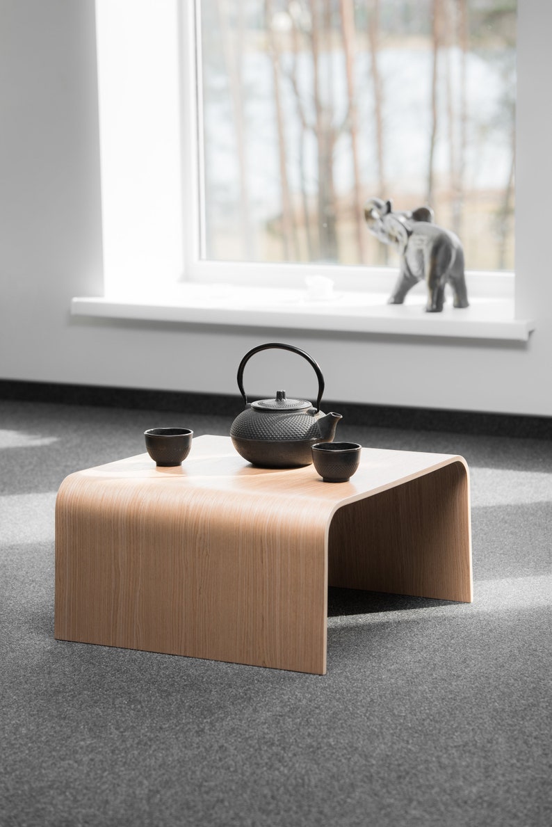 May include: A wooden table with a curved edge, featuring a black cast iron teapot and two small ceramic cups. The table is set on a gray carpet.