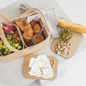 May include: A wooden picnic basket with a handle, filled with croissants, grapes, nuts, and a jar of jam. There are also two small wooden cutting boards, one with pistachios and the other with a piece of brie cheese.