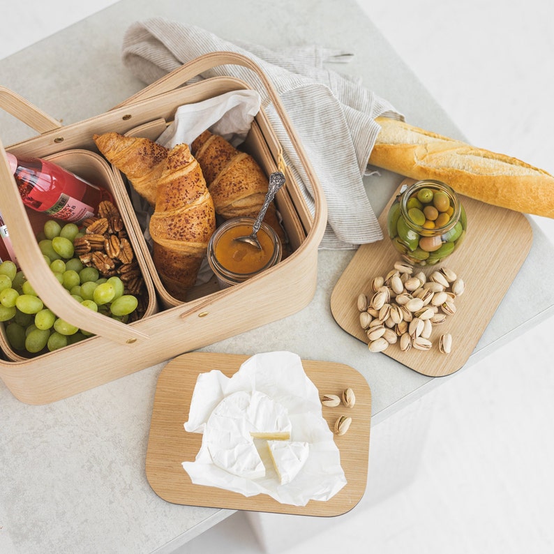 May include: A wooden picnic basket with a handle, filled with croissants, grapes, nuts, and a jar of jam. There are also two small wooden cutting boards, one with pistachios and the other with a piece of brie cheese.