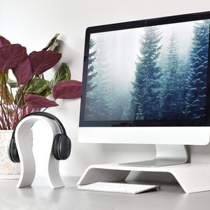 May include: White computer monitor stand with a white headphone stand on the left side. The monitor has a foggy forest scene as the desktop background. A white keyboard is in front of the monitor.