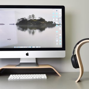 May include: A wooden headphone stand with a curved design holds a pair of black headphones. The stand is on a desk next to a silver computer monitor, a keyboard, and a wooden monitor stand.