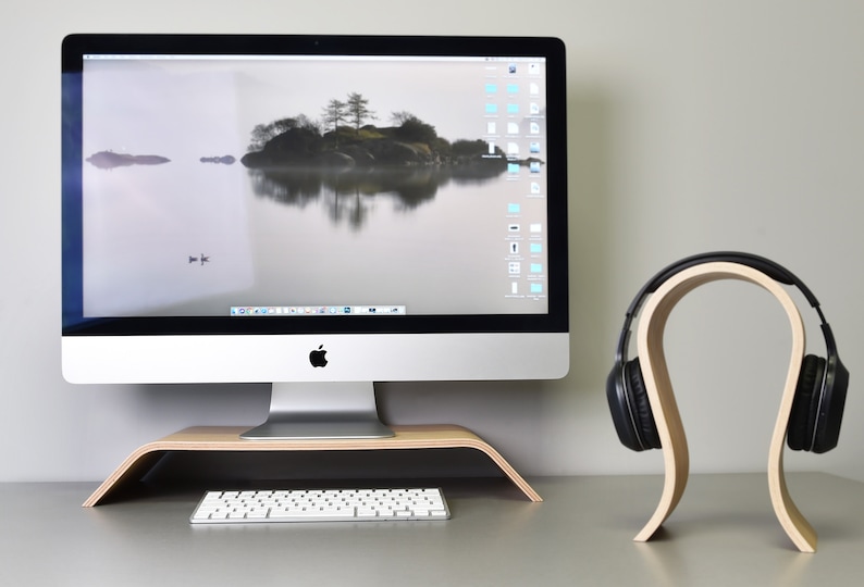 May include: A wooden headphone stand with a curved design holds a pair of black headphones. The stand is on a desk next to a silver computer monitor, a keyboard, and a wooden monitor stand.