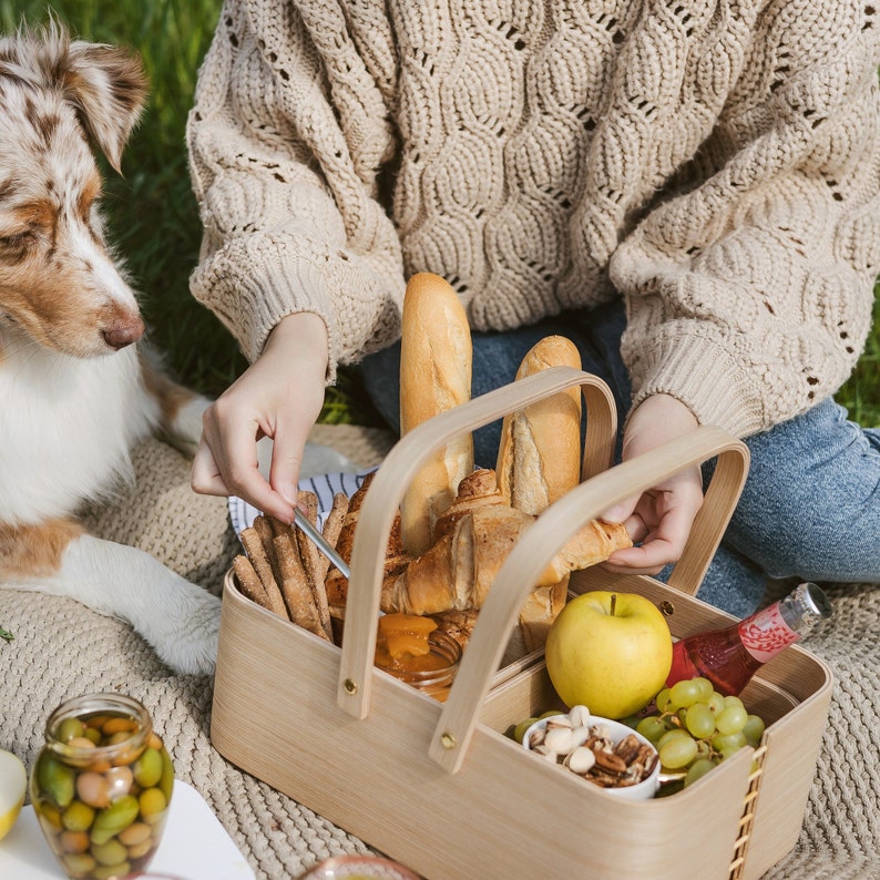 May include: A wooden picnic basket with a handle, filled with bread, fruit, and nuts. The basket is sitting on a blanket with a dog in the background.