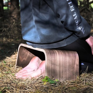 May include: A person sits on a wooden meditation stool with their feet resting inside the stool. The stool is made of dark wood with a natural grain pattern.