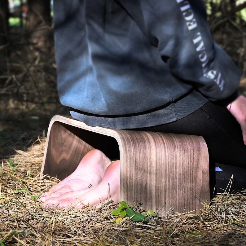 May include: A person sits on a wooden meditation stool with their feet resting inside the stool. The stool is made of dark wood with a natural grain pattern.