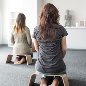 May include: Two people are sitting on wooden meditation cushions. The person in the foreground is wearing a gray t-shirt and blue jeans. The person in the background is wearing a beige sweater. Both people are sitting with their legs crossed.
