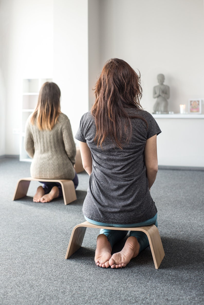 May include: Two people are sitting on wooden meditation cushions. The person in the foreground is wearing a gray t-shirt and blue jeans. The person in the background is wearing a beige sweater. Both people are sitting with their legs crossed.