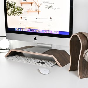 May include: A wooden headphone stand with a curved design, holding a pair of silver headphones. The stand is on a white desk with a computer monitor, keyboard, and mouse.