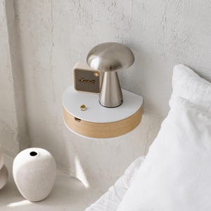 May include: A semi-circular wall-mounted floating shelf with a white top and a light wood grain drawer front. On the shelf sits a metallic mushroom-shaped lamp and a small beige speaker with the Marshall logo, plus a single gold ring.