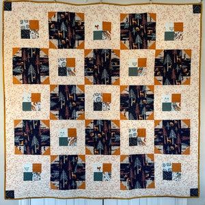 May include: A cream-colored quilt with a patchwork design featuring various shades of brown, black, and white. The quilt is made up of squares with different patterns, including trees, animals, and geometric shapes.