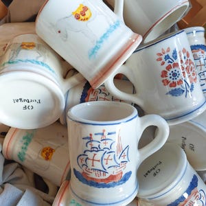 Op de afbeelding: Een verzameling witte keramische mokken met handgeschilderde ontwerpen. De ontwerpen tonen schepen, bloemen en paarden. De randen zijn rood of blauw en de bodems zijn gemarkeerd met "OF Portugal".