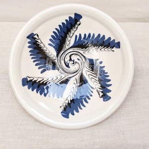 Puede incluir: Un plato de cerámica blanco con un diseño en espiral azul y negro en el centro. El diseño se asemeja a una flor o estrella estilizada. El plato tiene un borde ligeramente elevado.