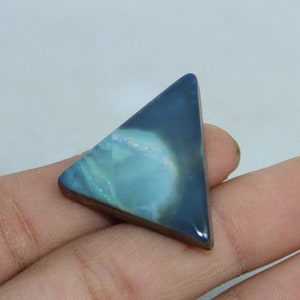 May include: A triangular opal gemstone with a dark blue and light blue color gradient. The stone has a polished surface and a wooden base. The opal displays iridescent flashes of color, and is held in a hand.