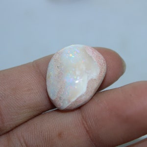 Puede incluir: Una piedra preciosa de ópalo pulido con una base blanca cremosa y destellos iridiscentes de color. La piedra tiene una forma redondeada y orgánica con una matriz rosada-marrón en los bordes. El ópalo se sostiene en una mano.