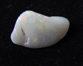 May include: A polished, oval-shaped opal gemstone. The opal is primarily white with iridescent flashes of red, green, and yellow. The stone has a smooth, glossy surface and a few small imperfections.