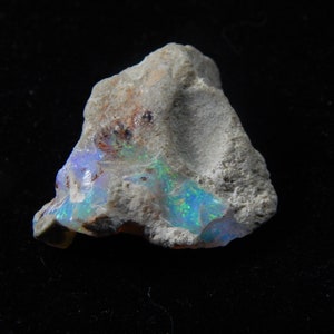 May include: A rough, natural opal gemstone with a light gray matrix. The opal displays iridescent flashes of blue, green, and yellow. The stone has a textured surface and irregular shape, showcasing its raw, unpolished beauty.