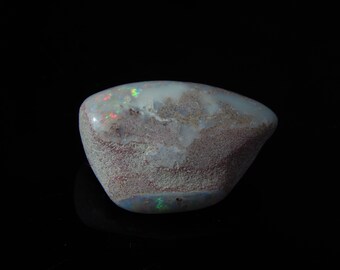 May include: A polished opal gemstone with a smooth, rounded shape. The stone features a mix of colors, including white, brown, red, and green, with a speckled texture. The opal is set against a black background.