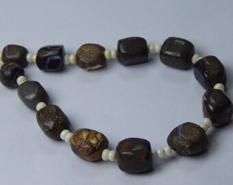 May include: A necklace featuring dark brown, irregularly shaped beads with cream-colored spacers. The beads have a mottled appearance with hints of blue and white, strung together to create a unique jewelry piece.