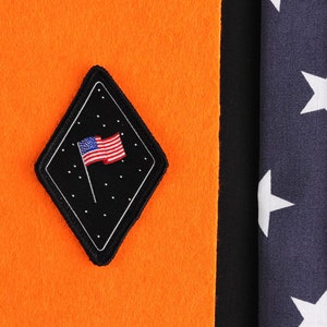 Peut inclure: Un écusson noir en forme de diamant avec un petit drapeau américain sur fond noir. L'écusson est cousu sur un fond de tissu orange. Le tissu est à côté d'un drapeau américain bleu et blanc.