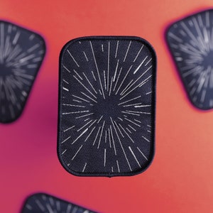 May include: A black patch with a white starburst design. The patch has a stitched border.