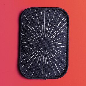 May include: Black fabric patch with a white starburst design. The patch has a stitched border.