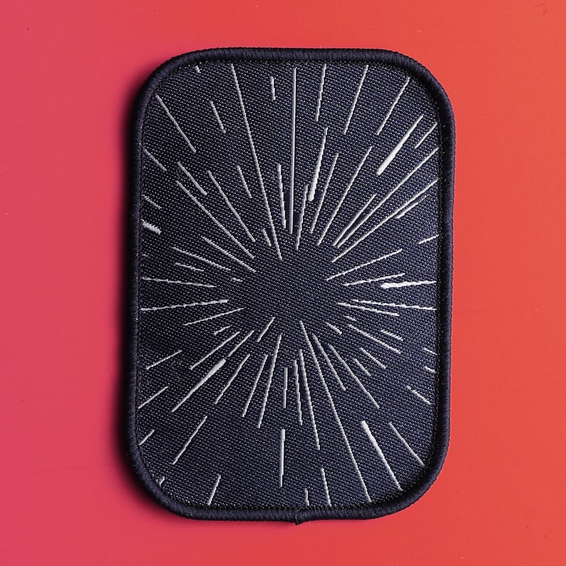 May include: Black fabric patch with a white starburst design. The patch has a stitched border.
