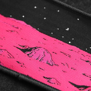 May include: A pink and black embroidered patch with a landscape design. The patch features a pink mountain range with a black sky with white stars.